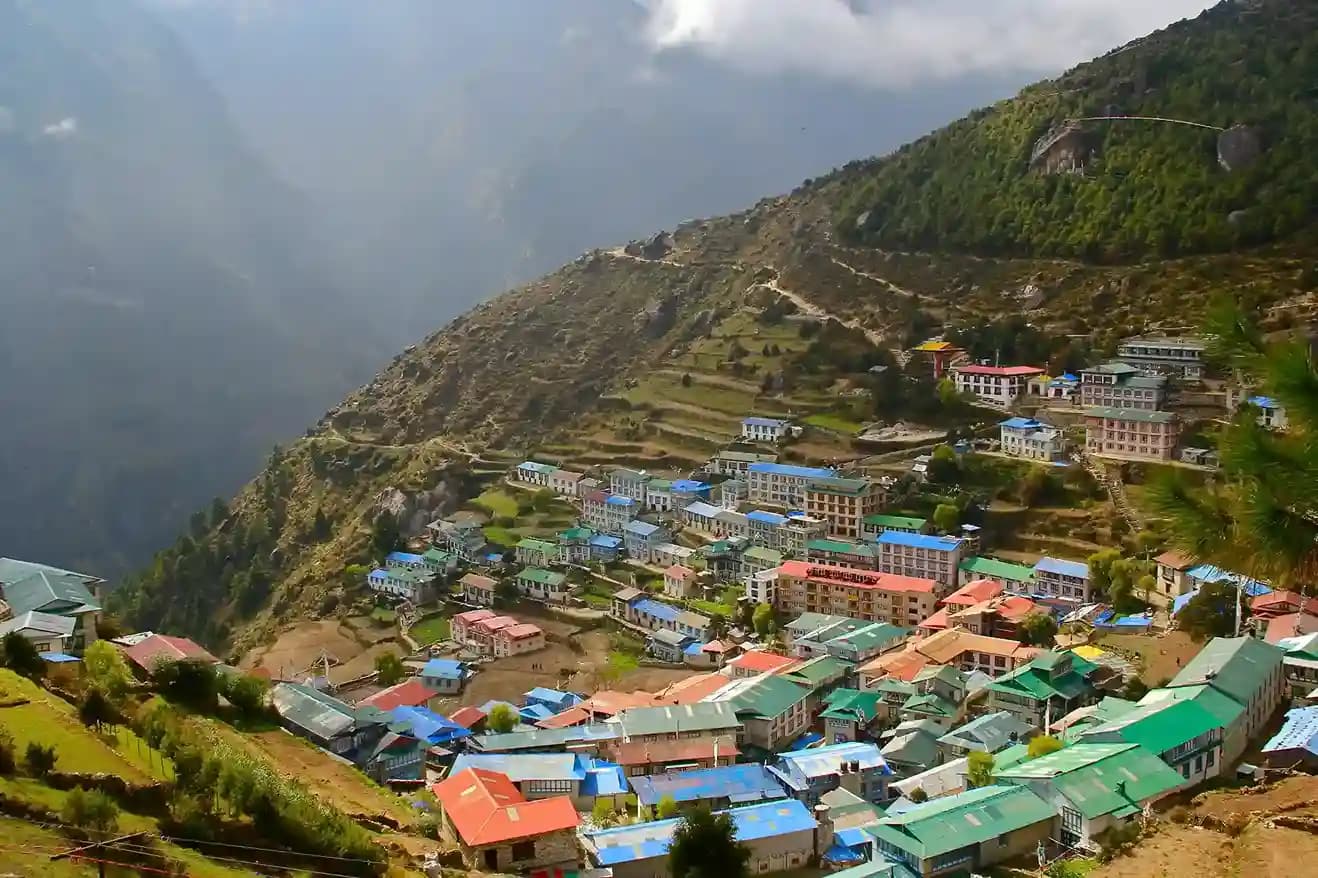 Aerial view of the horseshoe-shaped town of Namche Bazaar nestled in the mountainside with snow-capped peaks in the distance.